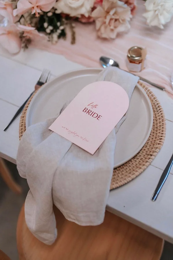 Pink arch wedding dual menu-place card sitting upon a table setting of a linen napkin and rattan placemat. Surrounded by flowers and a pink table runner.