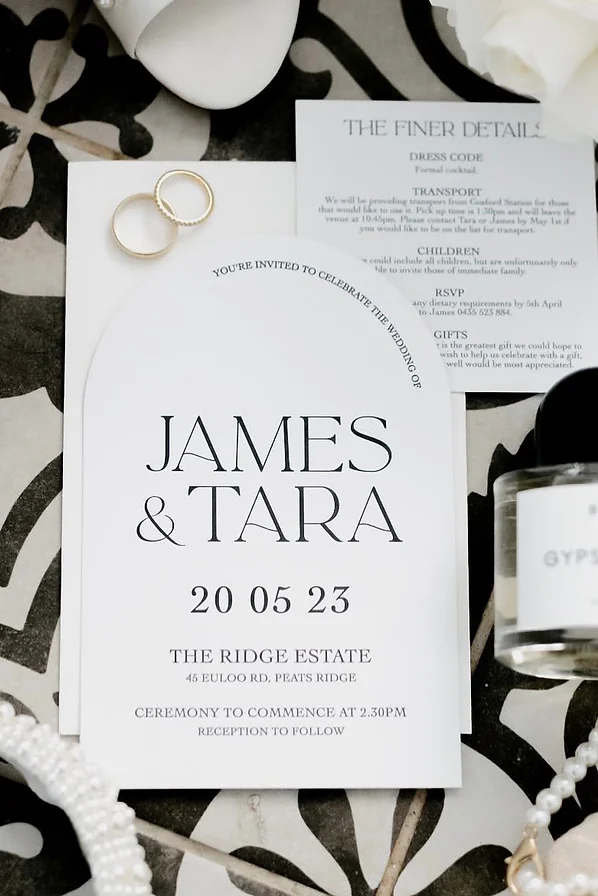 White arch letterpress invitation sitting as a flatlay for wedding photography with perfume and wedding rings.