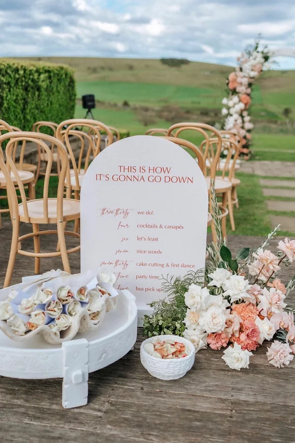 Order of events wedding signage in pink arch with florals and ceremony setup in the background.