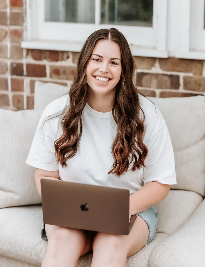 Liv from Gracie Creative sitting on a lounge with a laptop on her lap, smiling at the camera.