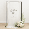 Linen wedding welcome sign hanging on a black metal frame with white ribbon, featuring black text for couple Brittany and Ryan, styled with white roses and greenery at the base – semi-custom signage by Gracie Creative, Australia.