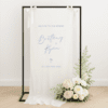 Linen wedding welcome sign hanging on a black metal frame with white ribbon, featuring blue text for couple Brittany and Ryan, styled with white roses and greenery at the base – semi-custom signage by Gracie Creative, Australia.