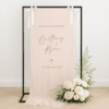 Linen wedding welcome sign hanging on a black metal frame with white ribbon, featuring brown text for couple Brittany and Ryan, styled with white roses and greenery at the base – semi-custom signage by Gracie Creative, Australia.