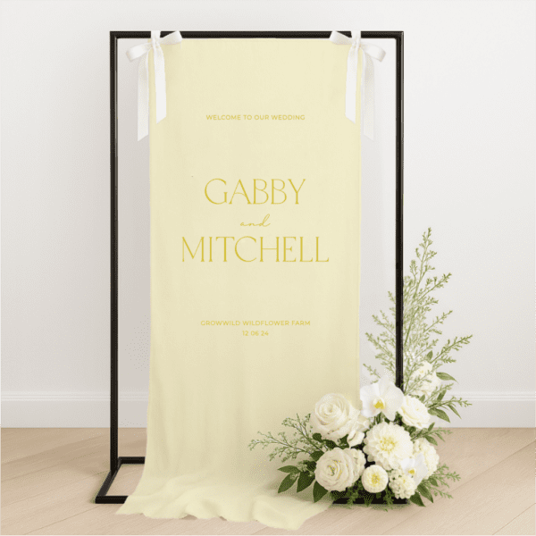 Linen wedding welcome sign hanging on a black metal frame with white ribbon, featuring yellow text for couple Gabby and Mitchell, styled with white roses and greenery at the base – semi-custom signage by Gracie Creative, Australia.
