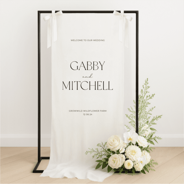 Linen wedding welcome sign hanging on a black metal frame with white ribbon, featuring black text for couple Gabby and Mitchell, styled with white roses and greenery at the base – semi-custom signage by Gracie Creative, Australia.