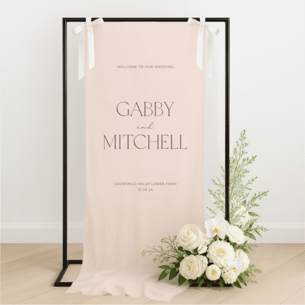 Linen wedding welcome sign hanging on a black metal frame with white ribbon, featuring merlot text for couple Gabby and Mitchell, styled with white roses and greenery at the base – semi-custom signage by Gracie Creative, Australia.